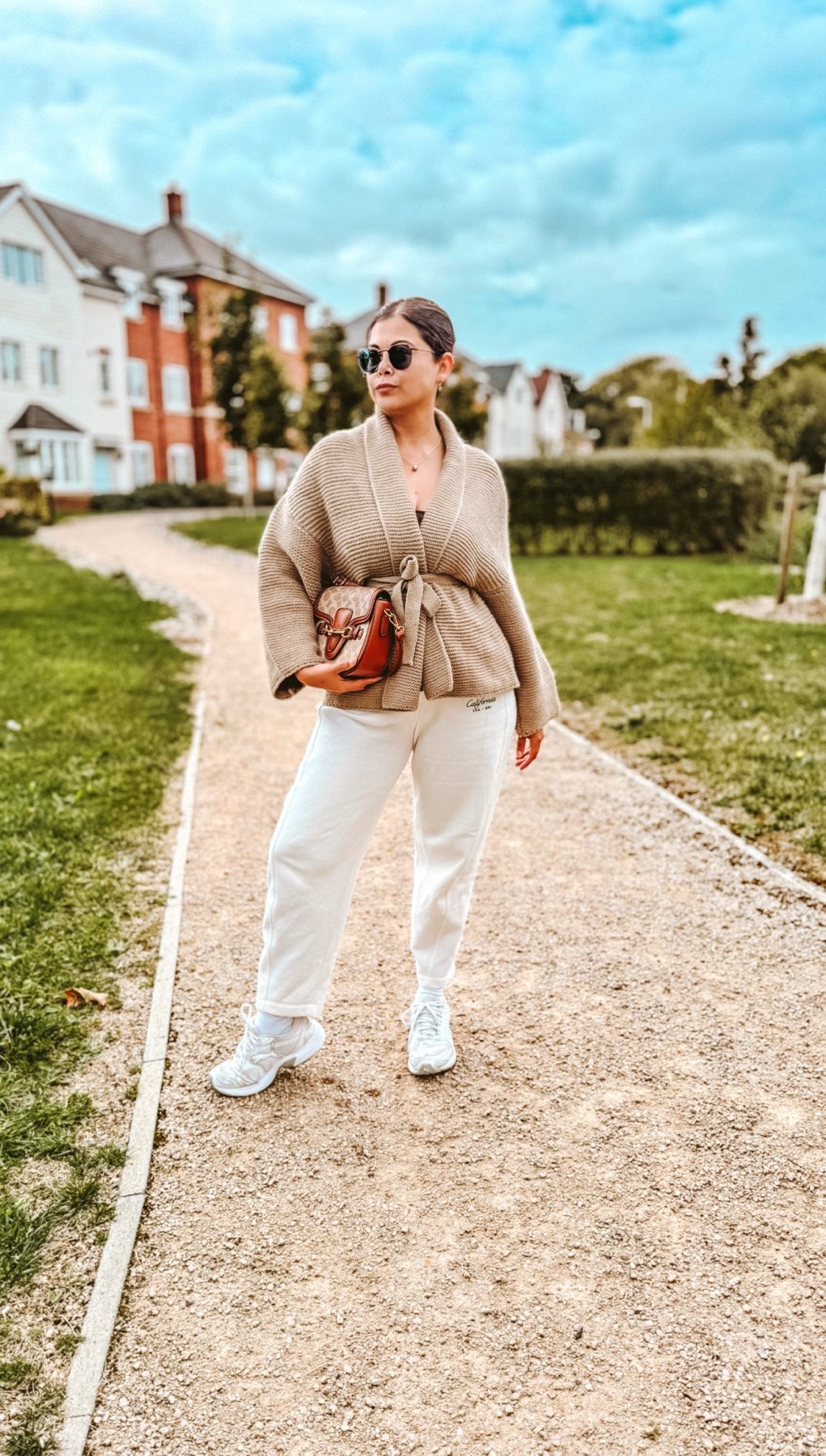 A woman in a cosy Autumn outfit with a beige wrap cardigan, white sweatpants , white sneakers and lovely coach bag in brown. Autumn outfits 