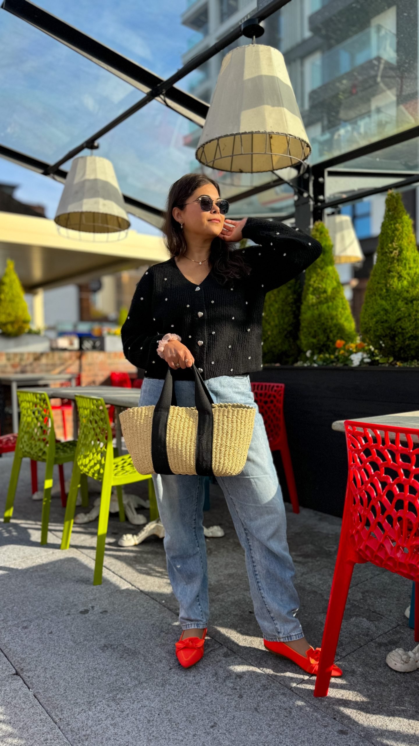 A girl wearing a black gemstone cardigan with heart buttons in black with her blue jeans and orange loafers. She is looking away from the camera smiling at the sun. 