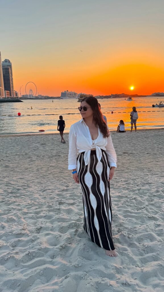 A woman standing on Palm Jumeirah  beach wearing a white h&M bikini set with a white shirt as a coverup and a black and white crochet knit maxi skirt the sun is setting behind her.  |Dubai beach outfit inspo
what to wear in Dubai | 