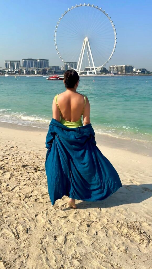 Dubai beachs| A woman walking towards the sea on JBR beach in Dubai wearing a green swimsuit and peacock blue beach coverup.