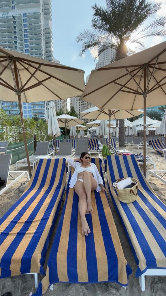 Dubai beach clubs| A woman sitting on a sunbed in a beach club in Dubai JBR beach wearing a white bikini.