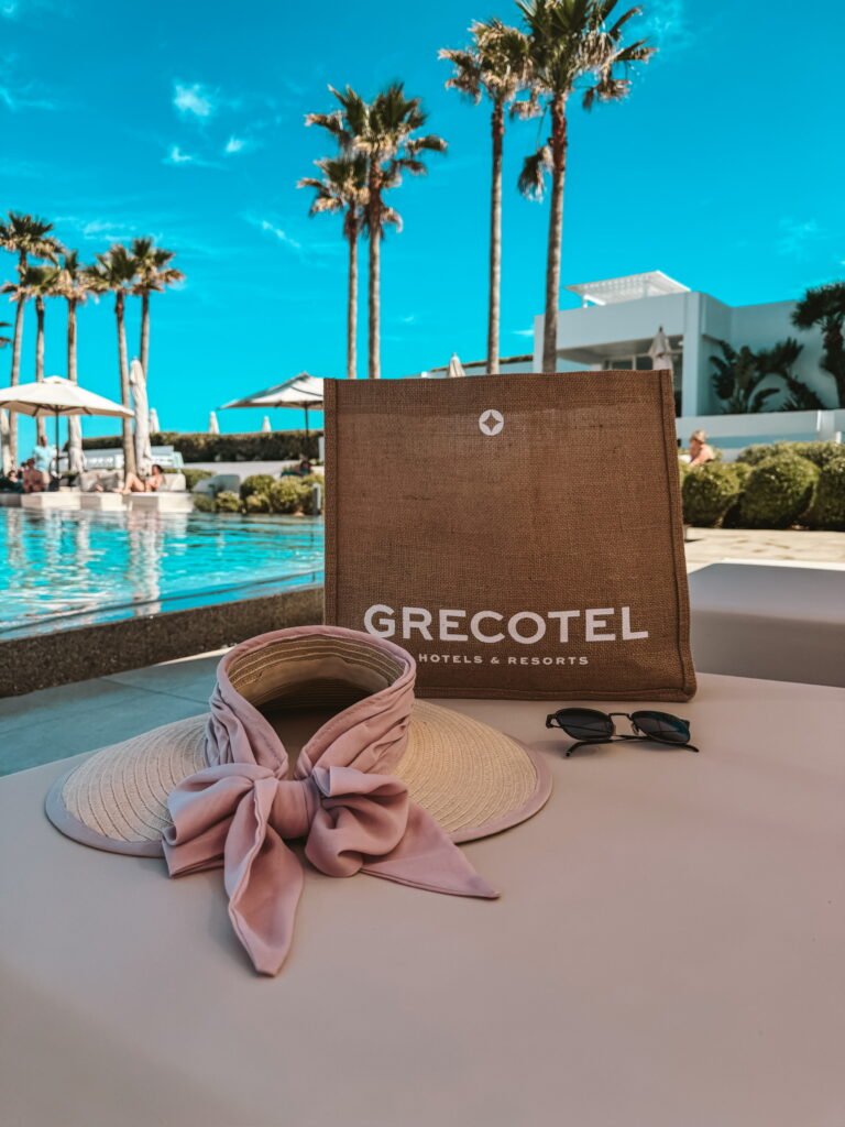 Beautiful straw hat with pink bow in the back on a sunbed by the pool with a beach bag and sunglasses in the background.