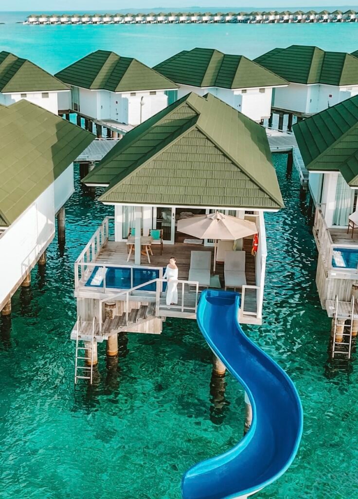 A drone shot of a woman in a white dress standing on the deck of Siyam World Water villa with pool and slide going into the ocean.