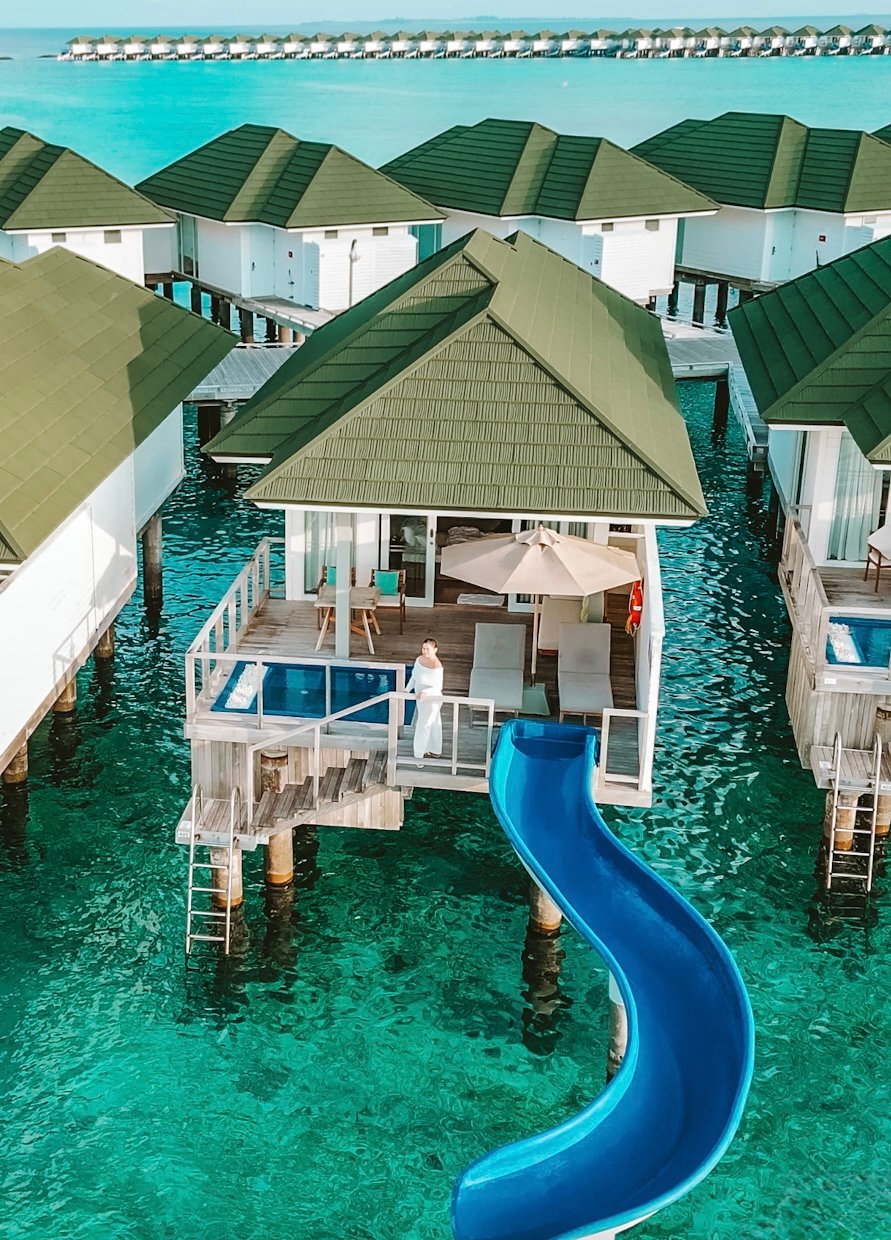 A drone shot of a woman in a white dress standing on the deck of Siyam World Water villa with pool and slide going into the ocean.