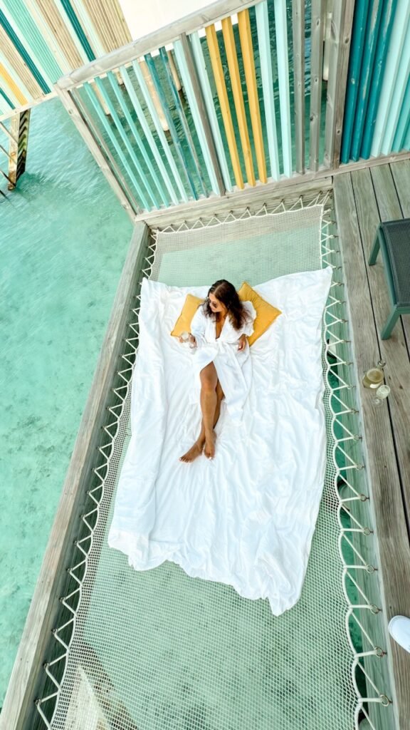 A woman sitting on a hammock on a watervilla in Hard Rock Maldives. She is wearing a bathrobe and drinking Champagne
