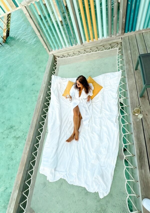 A woman sitting on a hammock on a watervilla in Hard Rock Maldives. She is wearing a bathrobe and drinking Champagne