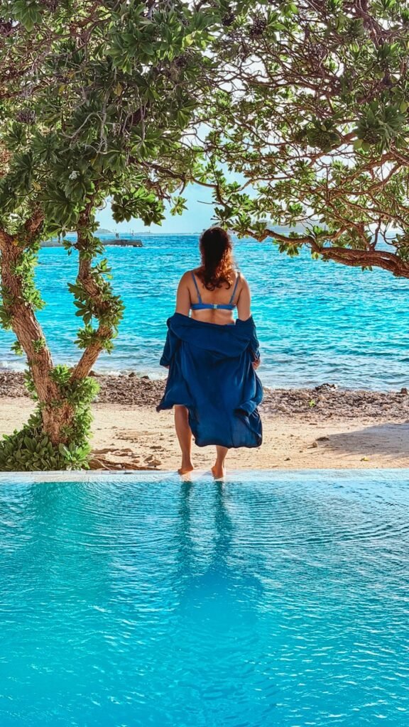 A woman in a blue bikini and a blue cover up standing at the edge of the infinity pool looking out to the sea in the Maldives.